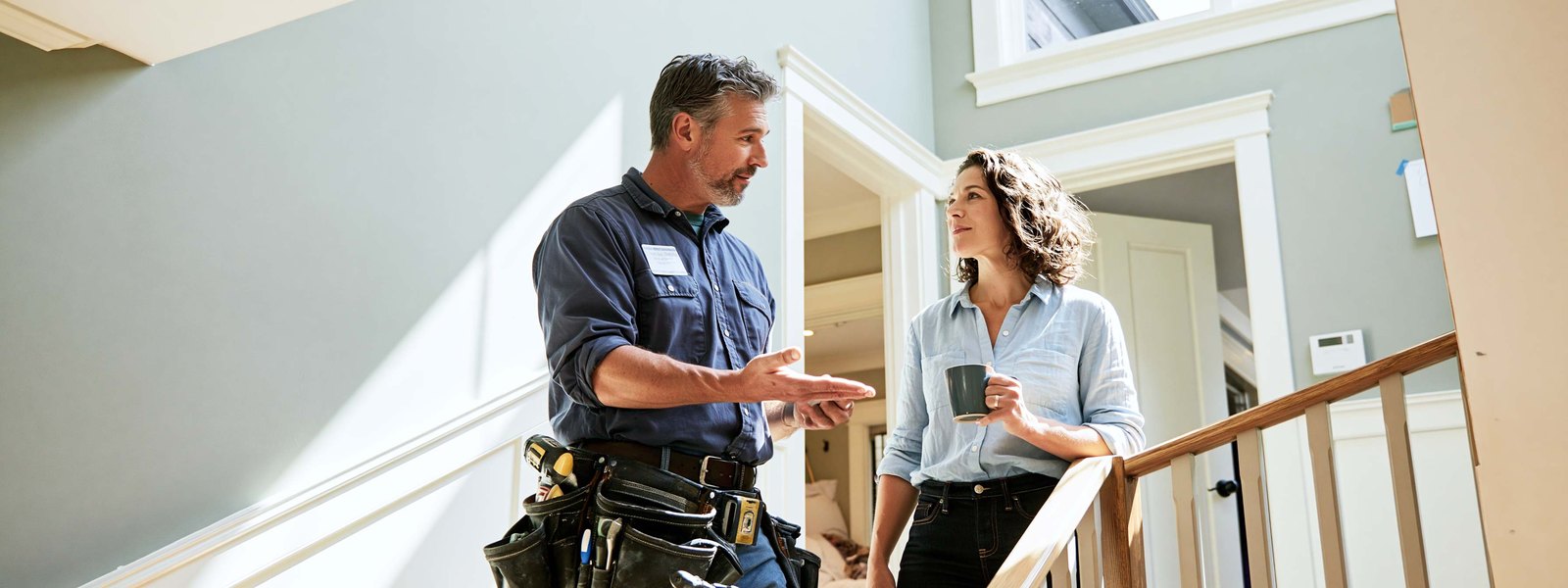 Drywall contractor in a work shirt and tool belt standing a few feet back from a long knockdown textured living room wall, one hand on the back of an armchair, looking along the surface toward a tall window
