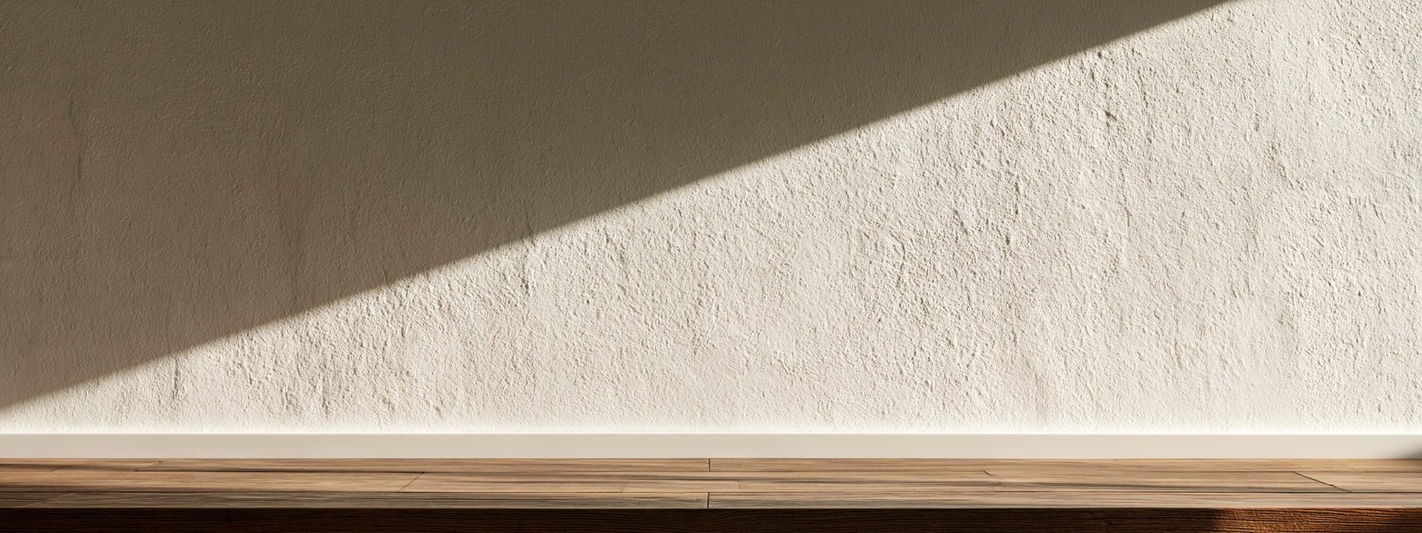 Long interior wall finished in knockdown and painted warm off white, late afternoon light raking across it from one side so the flattened plates throw soft shadow the whole length of the run