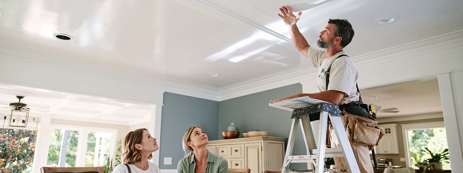 Drywall contractor on a stepladder in a furnished living room with one hand flat against the white ceiling above him, looking along the surface toward the far wall, a mud pan and a wide knife on the ladder shelf