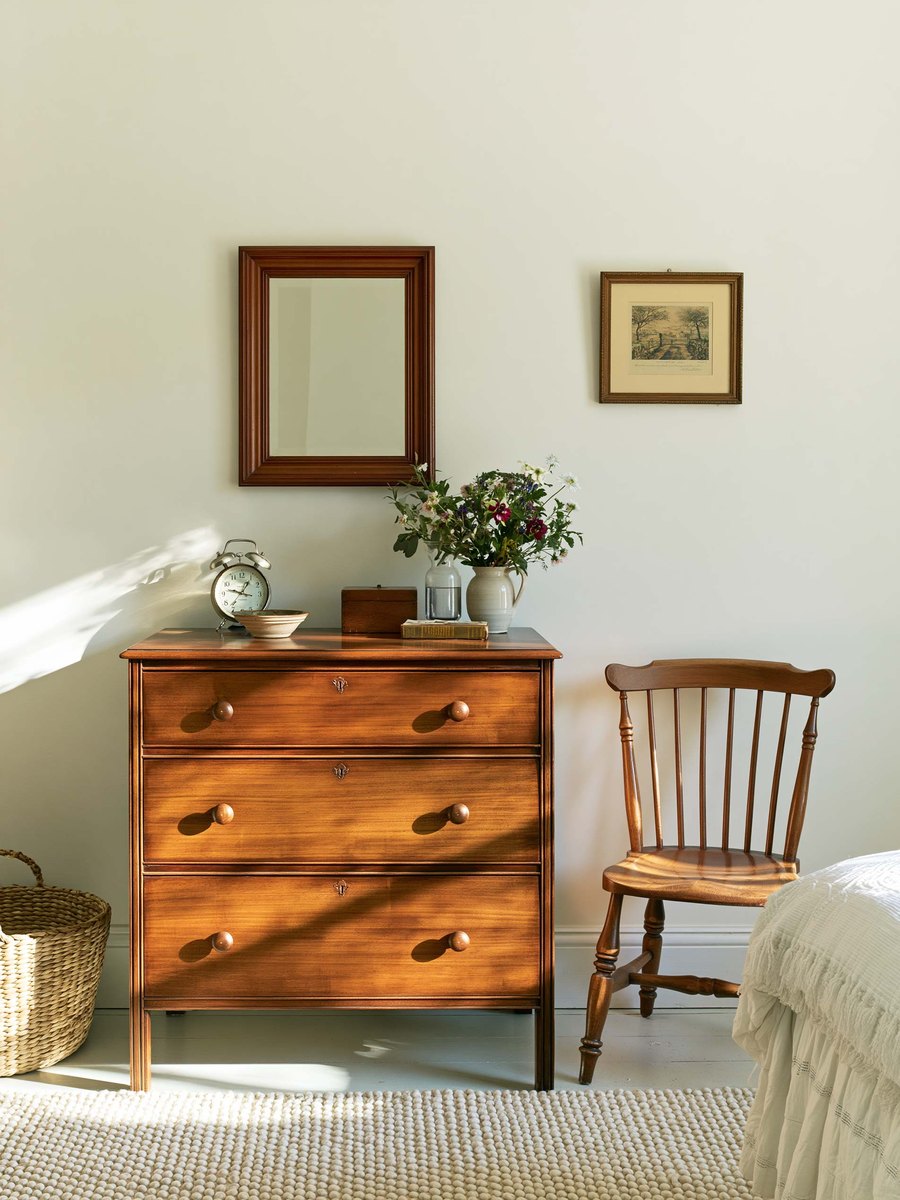 Homeowner holding a coffee mug in her own living room, a few steps from a long wall painted warm off white, looking along it in late afternoon light with a sofa and a floor lamp behind her
