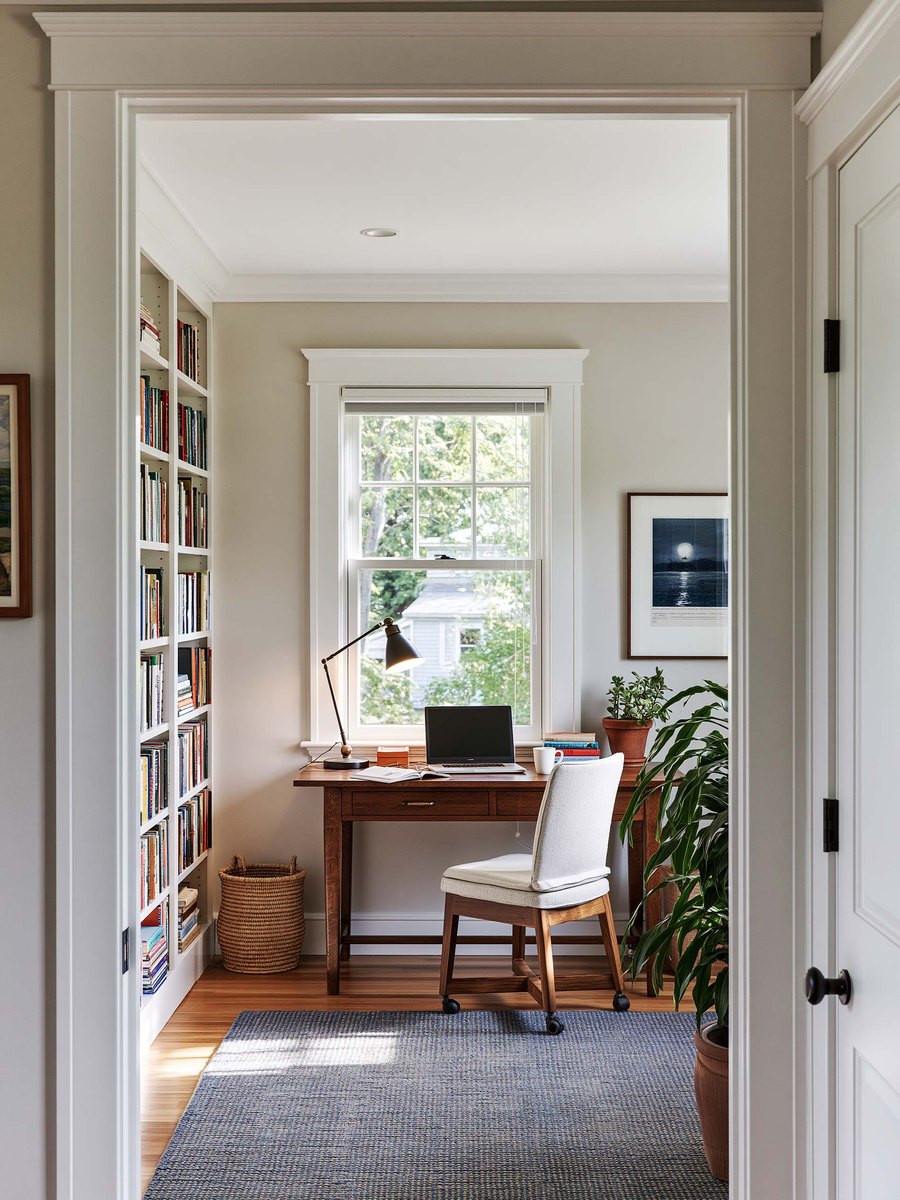 Homeowner standing in a bedroom doorway looking up at a flat white ceiling above a made bed, warm daylight coming in through a window at the far side of the room