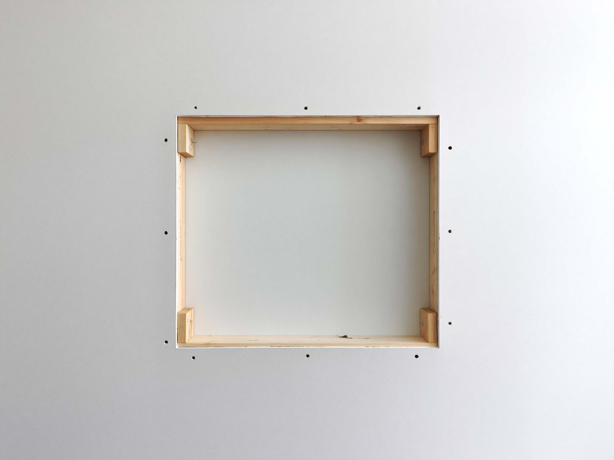 Knockdown textured living room wall cut open in a clean straight rectangle back to two studs, the cavity behind it dry and empty, the removed piece of board standing against the baseboard beside a lidded bucket and a mud pan on a canvas drop cloth
