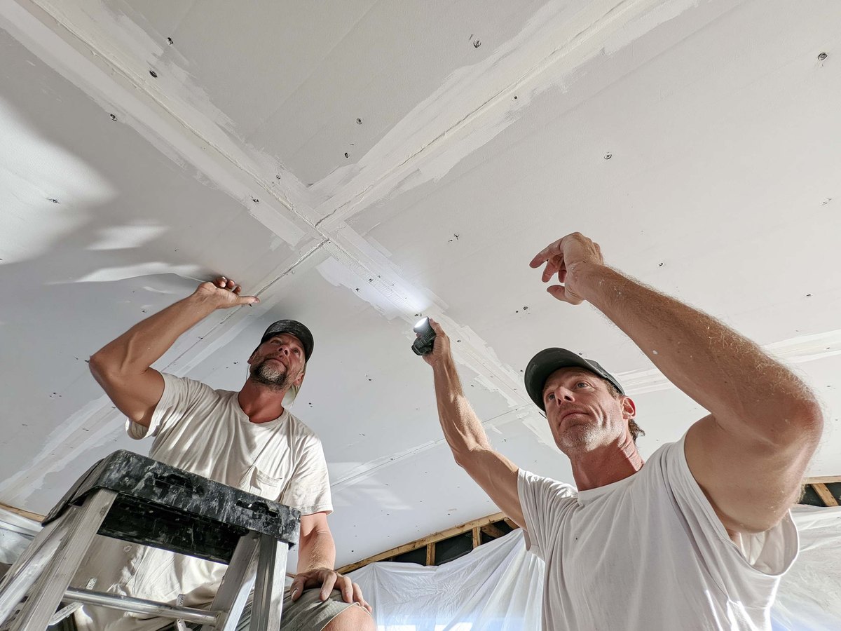 Bare ceiling seen from below after the old finish has been taken off, the taped joints and filled screw lines showing as pale bands across the board, a wide knife and a mud pan on a cloth-covered floor beneath