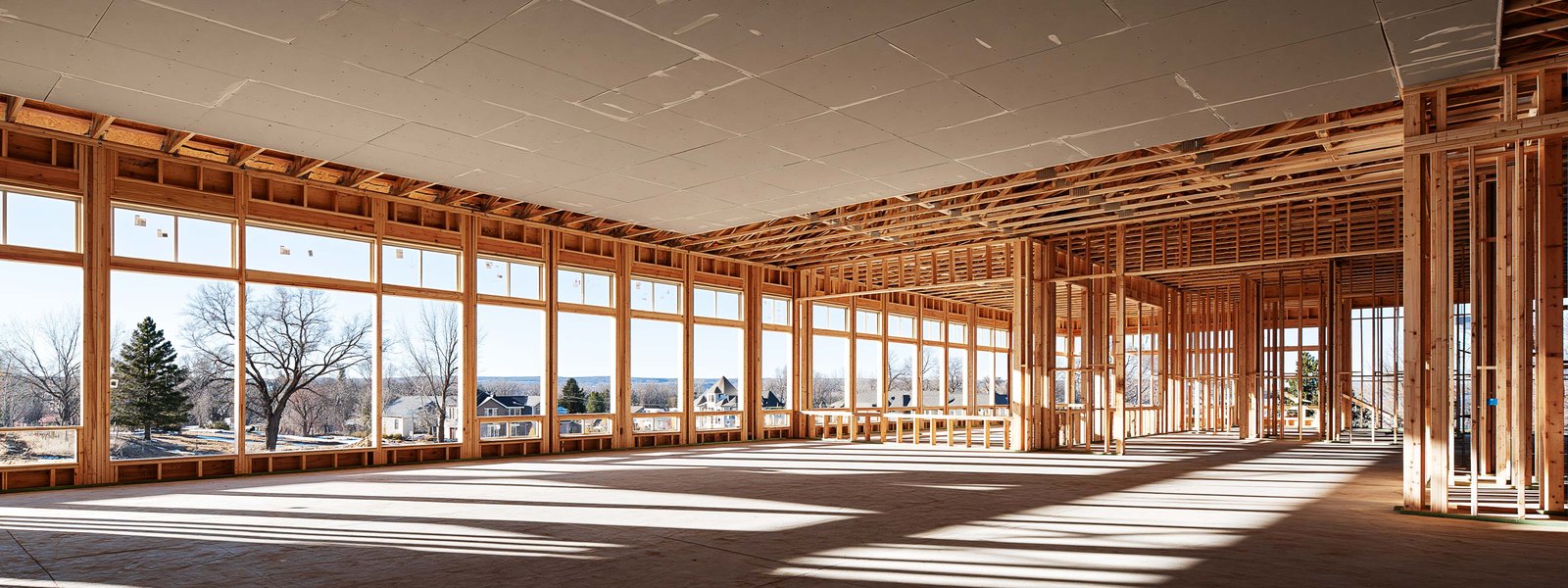 Interior of a house under construction with pale gray board hung across the studs of two rooms and a hallway, the joints open, daylight coming through an unglazed opening and the slab bare and swept
