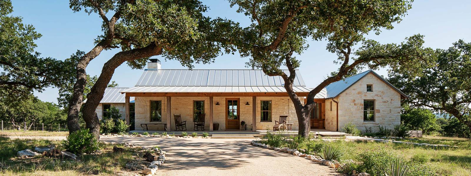 Single story house faced in buff limestone under a low shingled roof, a covered porch across the front between tall windows, live oaks arching over a gravel drive and open sky above the ridge behind