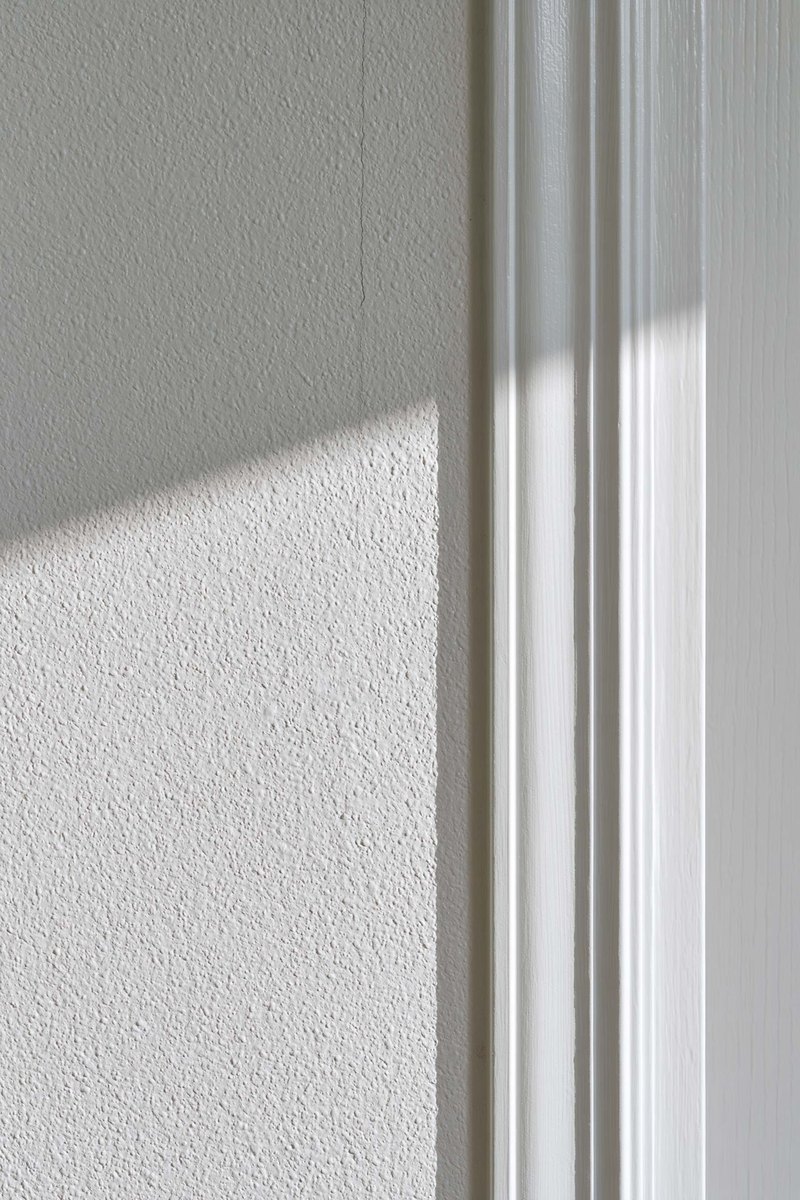 Hairline crack running up a painted interior wall from the top corner of a door frame, a short pencil tick marked beside it, low sun raking across the texture from the left